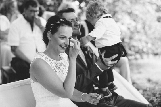 Hochzeitsfoto, Trauung, Braut wischt sich eine Träne ab, lächelt dabei. Im Hintergrund Ehemann und Kind zu sehen.